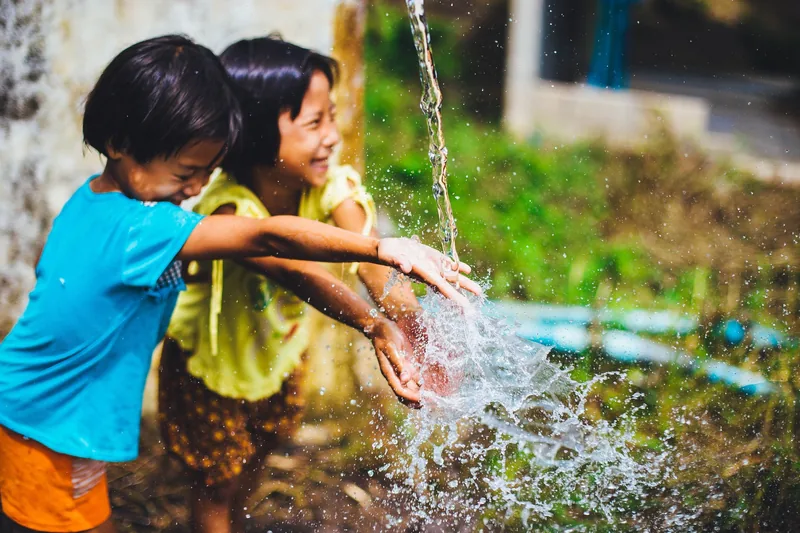 Two children laugh while playing with splashing water outdoors in Chiang Mai, Thailand.