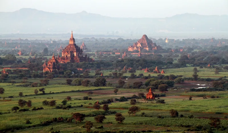 Ancient brick temples rise above a green plain in Bagan, Myanmar, seen from above at dawn.