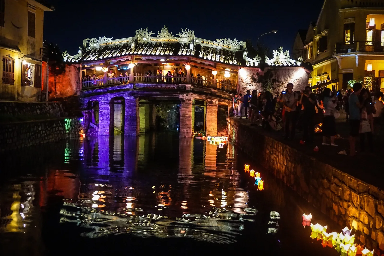 The Japanese Covered Bridge in Hoi An glows purple at night, reflected in the river with floating lanterns below.