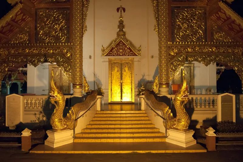 Golden naga serpent statues flank the illuminated entrance staircase of a Buddhist temple in Chiang Mai at night.